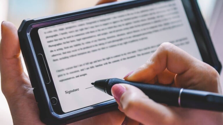 Hand hält Smartphone mit vergrößertem Textdokument auf dem Bildschirm, andere Hand hält Stift und bereitet sich darauf vor, auf der digitalen Unterschriftslinie zu unterschreiben.
