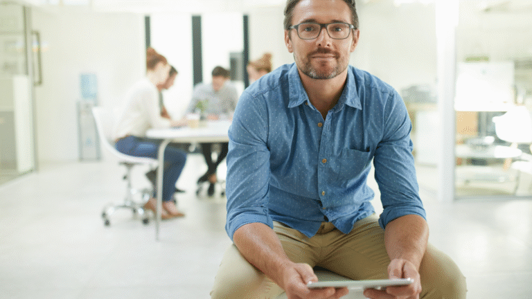 Ein Mann mit Brille und Bart trägt ein blaues Hemd und hält ein Tablet, während er in einem modernen Büro sitzt, im Hintergrund arbeiten drei Personen an einem Tisch.