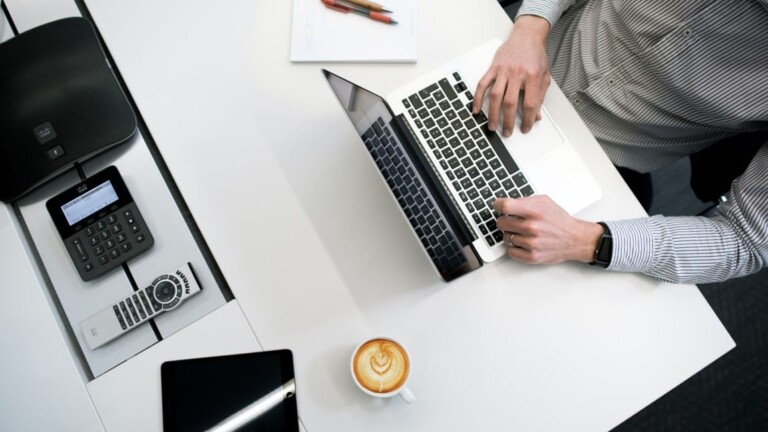 Von oben fotografierter weißer Schreibtisch mit einer Person in gestreiftem Hemd, die einen Laptop bedient, daneben eine Tasse Latte Macchiato mit Milchschaumherz, ein schwarzes Tablet, ein Telefon, eine Fernbedienung und ein Notizblock mit Stiften.