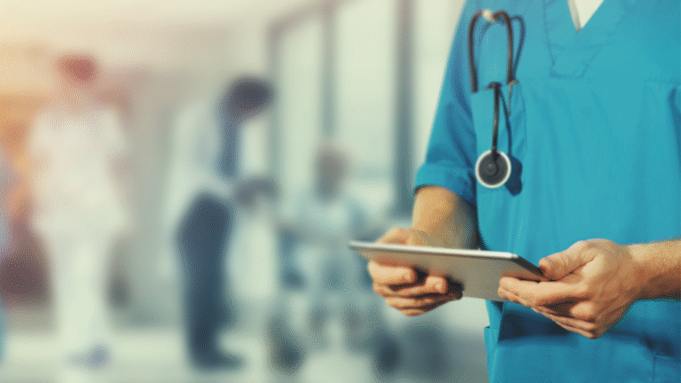 Arzt oder medizinisches Fachpersonal in blauer Schutzkleidung mit Stethoskop um den Hals hält ein Tablet, im Hintergrund unscharfe Szene mit weiteren medizinischen Personen und einem Patienten im Rollstuhl in einem hellen Krankenhausflur