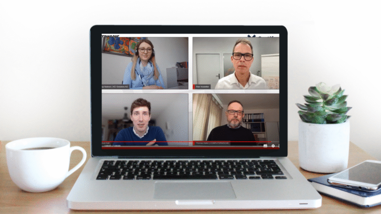 Laptop auf Holztisch zeigt Videokonferenz mit vier Teilnehmern, links neben dem Laptop eine weiße Kaffeetasse, rechts ein Topf mit Sukkulente und zwei übereinanderliegende Notizbücher mit Smartphone darauf