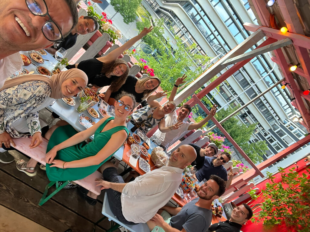 Gruppenfoto von elf fröhlichen Menschen, die an einem gedeckten Tisch auf einer Terrasse mit Blumen und moderner Architektur im Hintergrund sitzen, einige machen Friedenszeichen und lächeln in die Kamera.