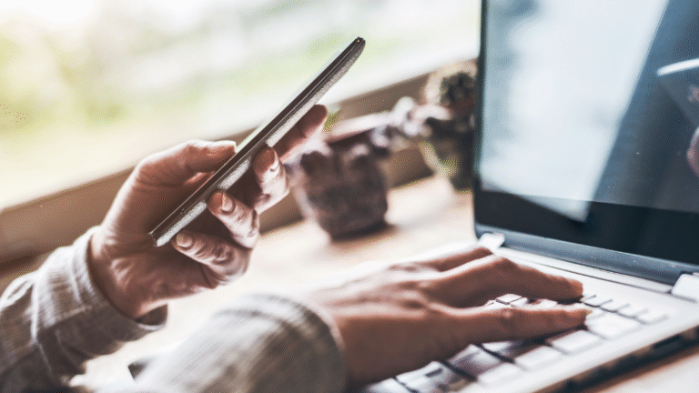 Nahaufnahme von Händen, die ein Smartphone halten und gleichzeitig auf der Tastatur eines Laptops tippen, mit unscharfem Hintergrund eines Fensters und Topfpflanzen auf einem Tisch.