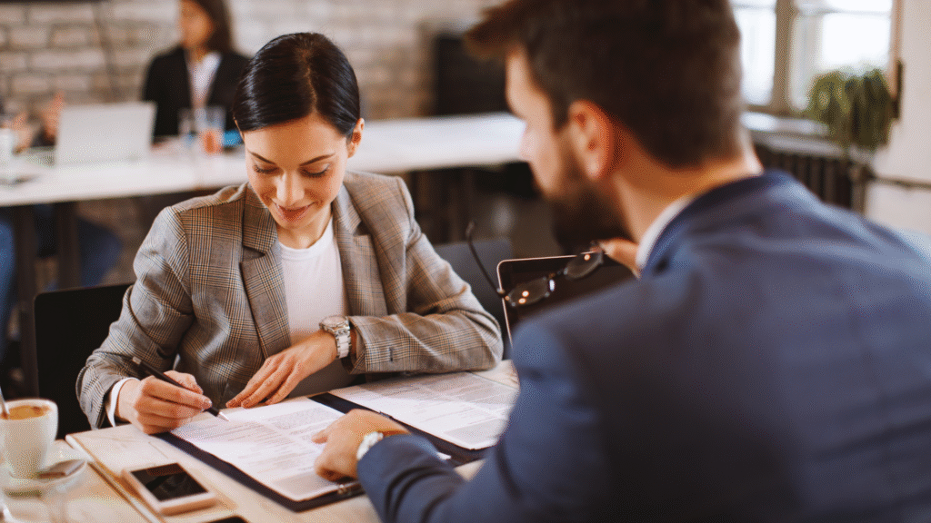 Woman in a checked blazer signs documents at a table while a man in a suit sits opposite her and points to the papers, office space with blurred people and plants in the background.