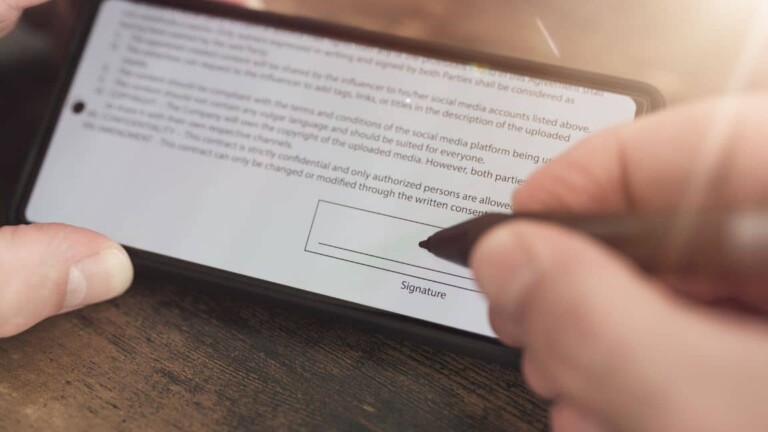 Hand unterschreibt digitale Vereinbarung auf einem Smartphone, das auf einem Holztisch liegt, mit einem Eingabestift.