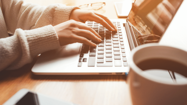 Hände in warmem Pullover, die auf einer Laptop-Tastatur tippen, daneben eine Tasse Kaffee und im Hintergrund eine Brille auf dem Tisch