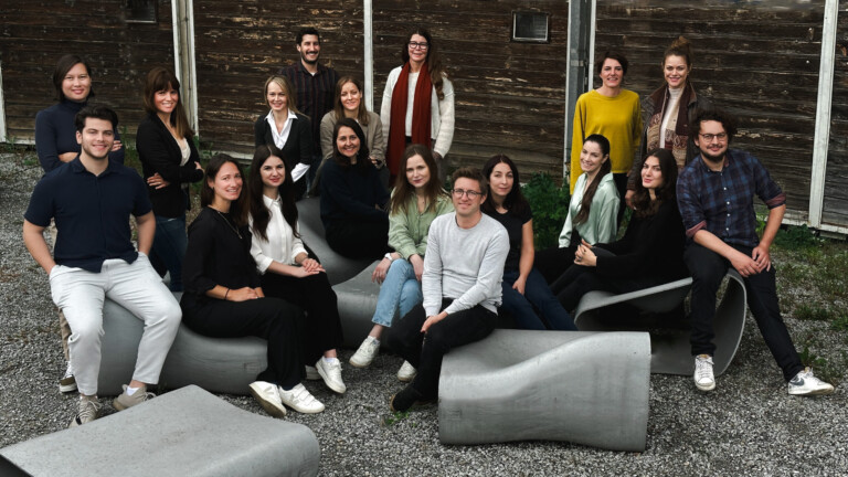 Gruppenfoto von 17 Personen, die vor einer rustikalen Holzwand im Freien auf modernen Betonbänken und dem Kiesboden sitzen und stehen, alle lächeln und in legere Kleidung gekleidet sind.
