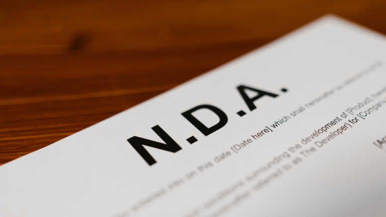 Nahaufnahme einer auf einem Holztisch liegenden, schräg fotografierten NDA-Vereinbarung mit hervorgehobenem fettem Text "N.D.A."