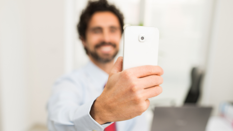 Ein Mann in Hemd und Krawatte hält ein weißes Smartphone in den Vordergrund und macht ein Selfie, während ein Büro mit Laptop unscharf im Hintergrund zu sehen ist.