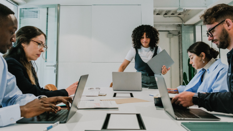 Fünf Personen in einem modernen Besprechungsraum: vier sitzen an beiden Seiten eines Tisches und arbeiten an Laptops mit Dokumenten und Notizen, in der Mitte steht eine Frau mit lockigem Haar und hält ein Tablet vor einem geöffneten Laptop.