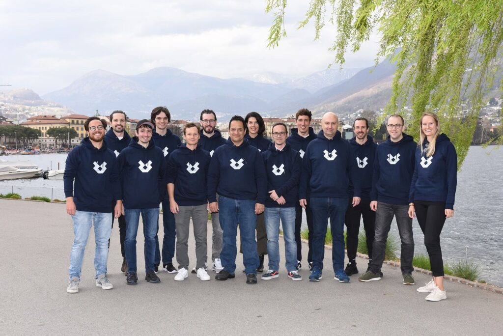 Gruppenbild von 14 Personen mit dunkelblauen Sweatshirts mit weißem Logo, posierend am Seeufer mit Bergen und Gebäuden im Hintergrund unter bewölktem Himmel