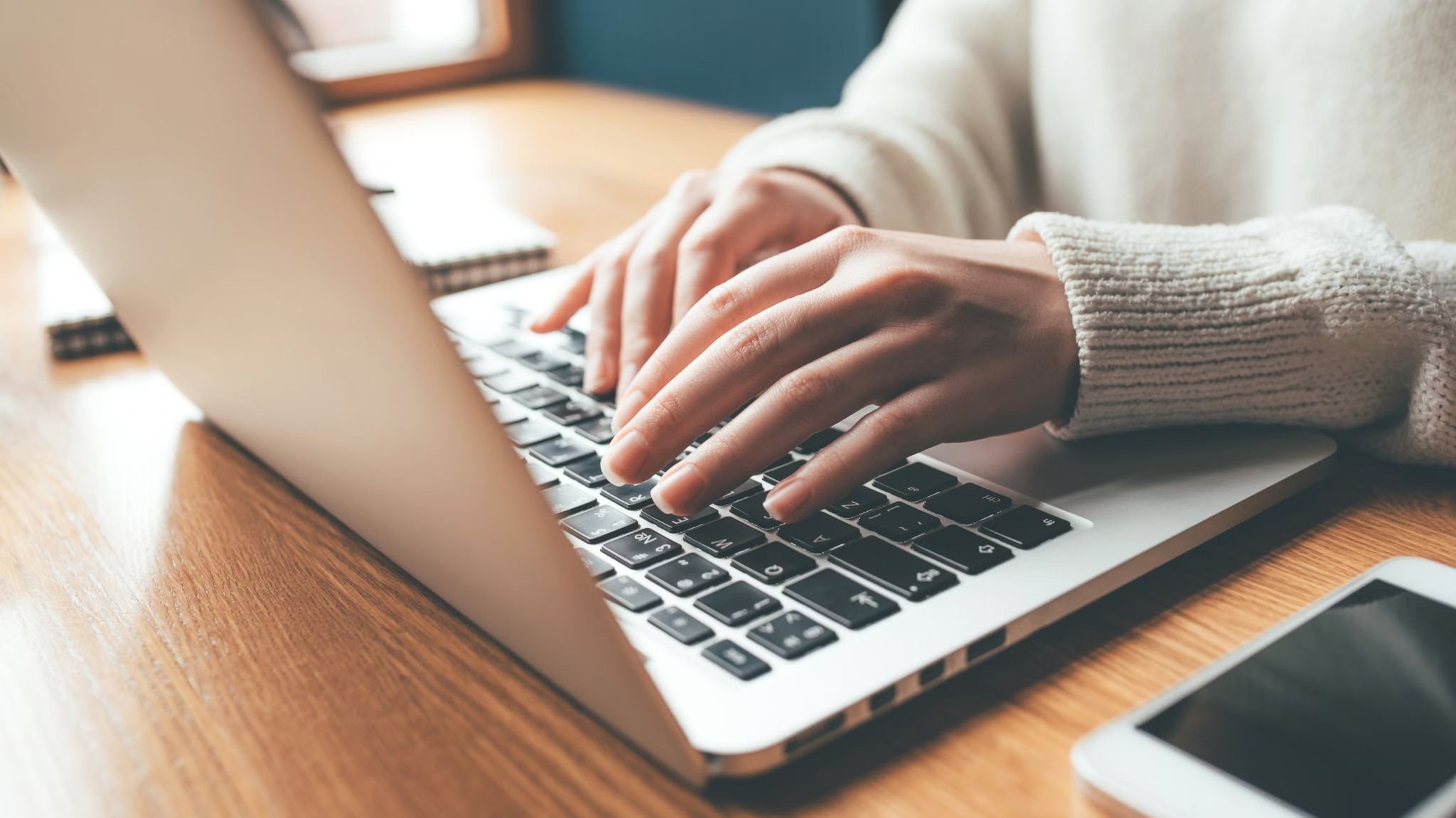 Hände in hellem Strickpullover tippen auf der Tastatur eines silbernen Laptops auf einem Holztisch; rechts liegt ein Smartphone und im Hintergrund sind Notizbücher sichtbar.